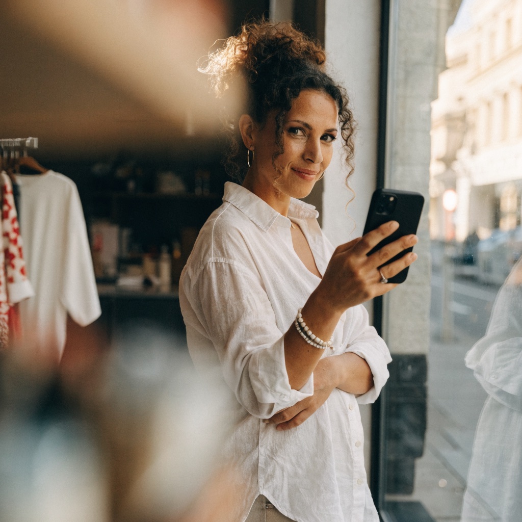 Shop owner standing at her store window, smiling while checking her phone